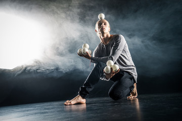 Wall Mural - Blond juggler sitting on the floor with white balls on black background