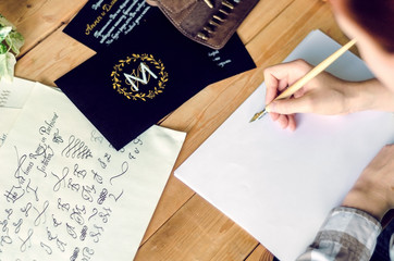 A girl calligrapher paints pen on white paper behind her working wooden table.