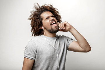 Wall Mural - A curly-headed handsome man wearing a gray T-shirt is listening to music in the earphones with an outrageous look over the white background.