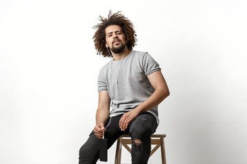 Wall Mural - A curly-headed handsome man wearing a gray T-shirt and ripped jeans is listening to music in the earphones and sitting on the backless stool over the white background.