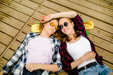 Two stylish young urban girls with longboards lie on the wooden flooring in the street. Friends have fun and spend time together in the summer during the holidays