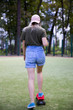 From the back, a young girl in short blue shorts, a T-shirt and a cap rides on a skateboard in the park in the summer
