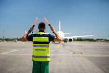 Directing The Jet. Back View Of Aviation Marshaller At Airport. Aircraft, Runway And Sky On Blurred Background