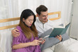 © bixpicture - Beautiful pregnant woman holding smart tablet computer and husband reading book on the bed in bedroom. Concept of couple, lover, family happy, lifestyle, relax and holiday.