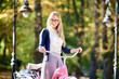 Portrait of young smiling happy attractive blond long-haired woman in glasses, skirt and blouse holding handles of pink lady bicycle on blurred colorful golden yellow green bokeh foliage background.