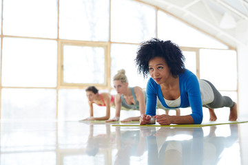 Diverse confident sportive women training abdomen with plank exercise on yoga mats in spacious white hall