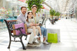 © pressmaster - Adult parents with charming girl sitting on bench in big light shopping mall with colorful paper bags and smiling at camera