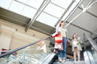 © pressmaster - From below shot of adult woman with little girl standing on moving stairs in shopping mall carrying paper bags