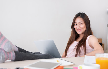 Poster - Asian female freelancer smile and holding laptop with rest leg on table in bedroom at home.Work at home concept.work from home concept.
