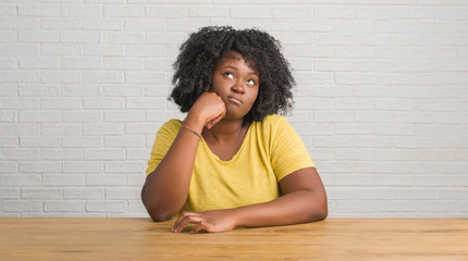 Wall Mural - Young african american woman sitting on the table at home with hand on chin thinking about question, pensive expression. Smiling with thoughtful face. Doubt concept.