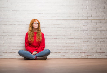 Wall Mural - Young redhead woman sitting over brick wall smiling looking side and staring away thinking.