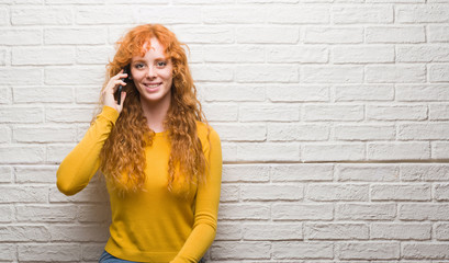 Wall Mural - Young redhead woman standing over brick wall talking on the phone with a happy face standing and smiling with a confident smile showing teeth