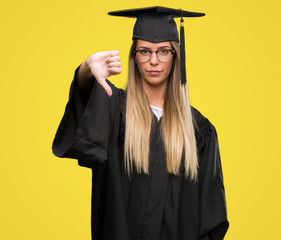 Wall Mural - Beautiful young woman wearing glasses and graduation robe with angry face, negative sign showing dislike with thumbs down, rejection concept