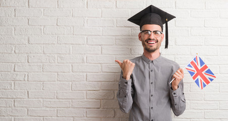 Young adult man over brick wall wearing graduation cap holding uk flag pointing and showing with thumb up to the side with happy face smiling