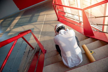 Engineer or Architect feeling tired and headache with his job. sitting on building stairs with architectural drawing on his side while wearing protective equipment safety helmet at construction site