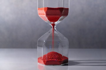 Red sand running through the shape of modern hourglass on white table.Time passing and running out of time. Urgency countdown timer for business deadline concept with copy space