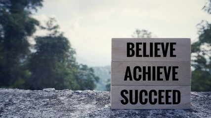 Wall Mural - Motivational and inspirational quote - ‘BELIEVE, ACHIEVE, SUCCEED’ written on wooden blocks. Blurred styled background.