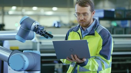 Fototapete - At the Factory: Automation Engineer Uses Laptop for Programming Robotic Arm. New Era in Automatic Manufacturing Industry.