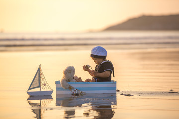 Wall Mural - Cute baby child, sweet boy, playing with boat, teddy bear and fishes on sunset at the edge of the ocean