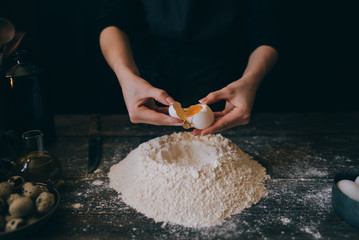 Woman cracking egg for dough. Retro styled imagery. Making dough. Cooking ingredients for pastry on rustic wood, culinary classes or recipe concept. Preparation of the dough from fresh ingredients.