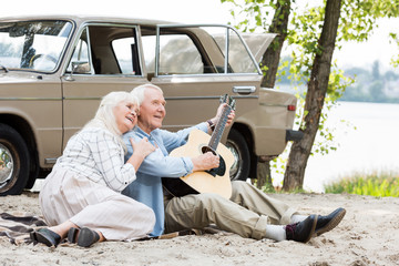 beautiful senior woman sitting on sand with man playing guitar against beige vintage car