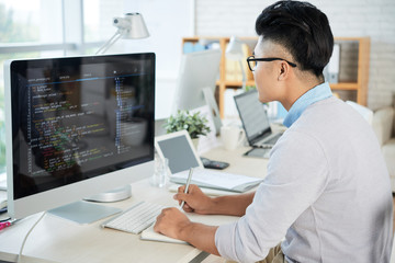 Side view of young Asian guy looking at computer monitor and drawing in sketchbook while working in modern office