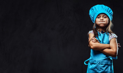 disgruntled little girl dressed in blue cook uniform standing with crossed arms. isolated on dark te