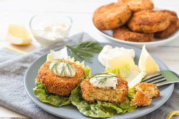 Salmon patties served on green lettuce leaves with fresh dill, lemon slices and white sauce on top.