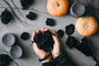 Witch's hands holding black roses over grey background surrounded black candles, small pumpkins, dried branches and vintage wineglass . Halloween concept, flat lay, top view.