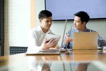 Two Asian coworkers sitting in an office looking at a digital tablet and laptop