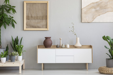 Real photo of a botanic living room interior with burlap artwork on the gray wall and white cupboard surrounded by plants