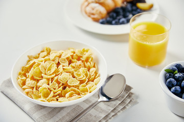 Wall Mural - close up view of corn flakes in bowl, glass of juice and croissant with blueberries and plum pieces for breakfast on white tabletop