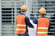 © DragonImages - Back view of men in hardhats and waistcoats holding draft and pointing at building while standing on construction site
