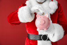 Santa Claus Putting Coin Into Piggy Bank On Color Background, Closeup