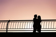 Young hugging couple silhouettes, standing near railing, on city embankment, against sunset sky.