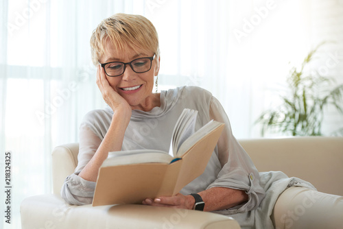 Cuadros en Lienzo Senior blond woman in glasses resting on sofa and reading a book