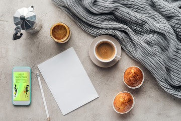 Wall Mural - top view of cup of coffee with muffins, blank paper and smartphone with best shopping app on screen on concrete surface with knitted wool drapery