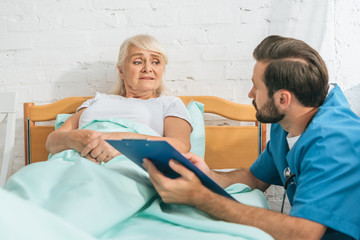 Wall Mural - male nurse writing on clipboard and looking at sick senior woman lying in hospital bed