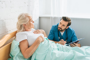 Wall Mural - doctor writing on clipboard and looking at sick senior woman lying in hospital bed