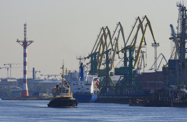 The tugboat sails in the roadstead of the seaport in the background is the pier with the port cranes and a lighthouse