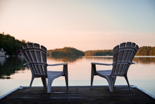 Muskoka Chairs Sitting At The End Of A Dock In Front Of Lake Joseph At Sunrise.