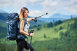 © anatoliy_gleb - Portrait of happy sporty woman tourist with blue backpack pointing away with her trekking sticks, smiling to the camera on summer day. Mountains, forests and cloudy sky on the blurred background