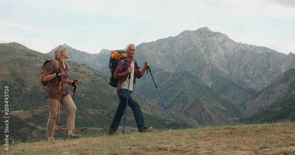 Active senior caucasian couple hiking in mountains with backpacks, enjoying their adventure 4k - obrazy, fototapety, plakaty 