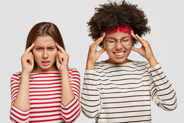 Wall Mural - Displeased young mixed race women keep fingers on temples, have headache after working on common project, look desperately at camera, stand against white background. Thoughtful multiethnic females