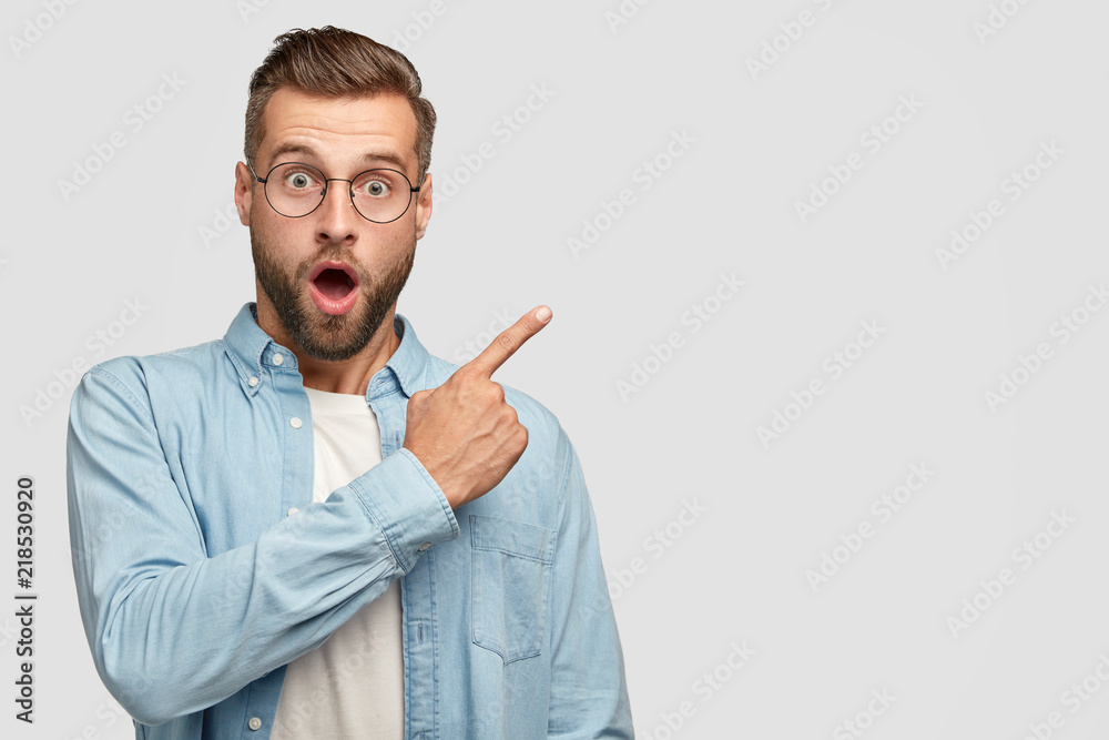 Stunned handsome unshaven man with shocked facial expression, dressed in fashionable shirt, points at upper right corner, shows blank space for your hearder, feels horrified. Advertisement concept - obrazy, fototapety, plakaty 