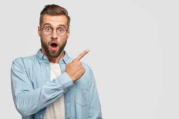 stunned handsome unshaven man with shocked facial expression, dressed in fashionable shirt, points a
