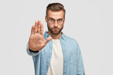 Serious European male shows stop gesture, demands something, has strict facial expression, wears round spectacles and formal shirt, isolated over white background. People and body language concept