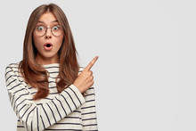 Horizontal Shot Of Beautiful Young Caucasian Female With Surprised Expression, Points At Upper Right Corner, Dressed In Striped Sweater, Wears Round Spectacles, Isolated Over White Background