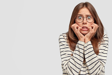 Wall Mural - Scared European young female looks with bugged eyes, keeps hands on cheeks, dressed in striped sweater, isolated over white background with copy space for your promotional content. Fear concept