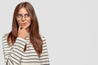 © WHstudio Leushin N - Confused lovely female teenager holds chin, looks thoughtfully aside, has dark hair, wears striped sweater, isolated over white background with copy space for advertisement. Thoughtful woman indoor